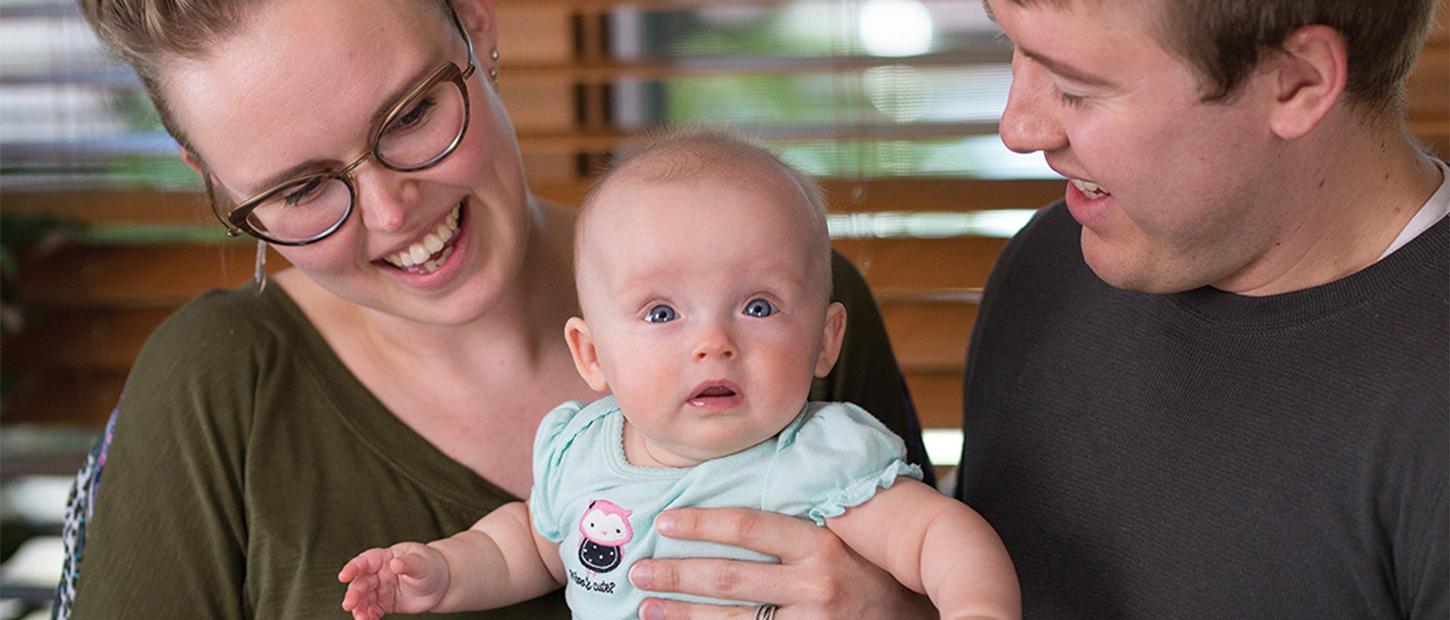 Mom and dad look happily at their baby while sitting together in front of a large window.