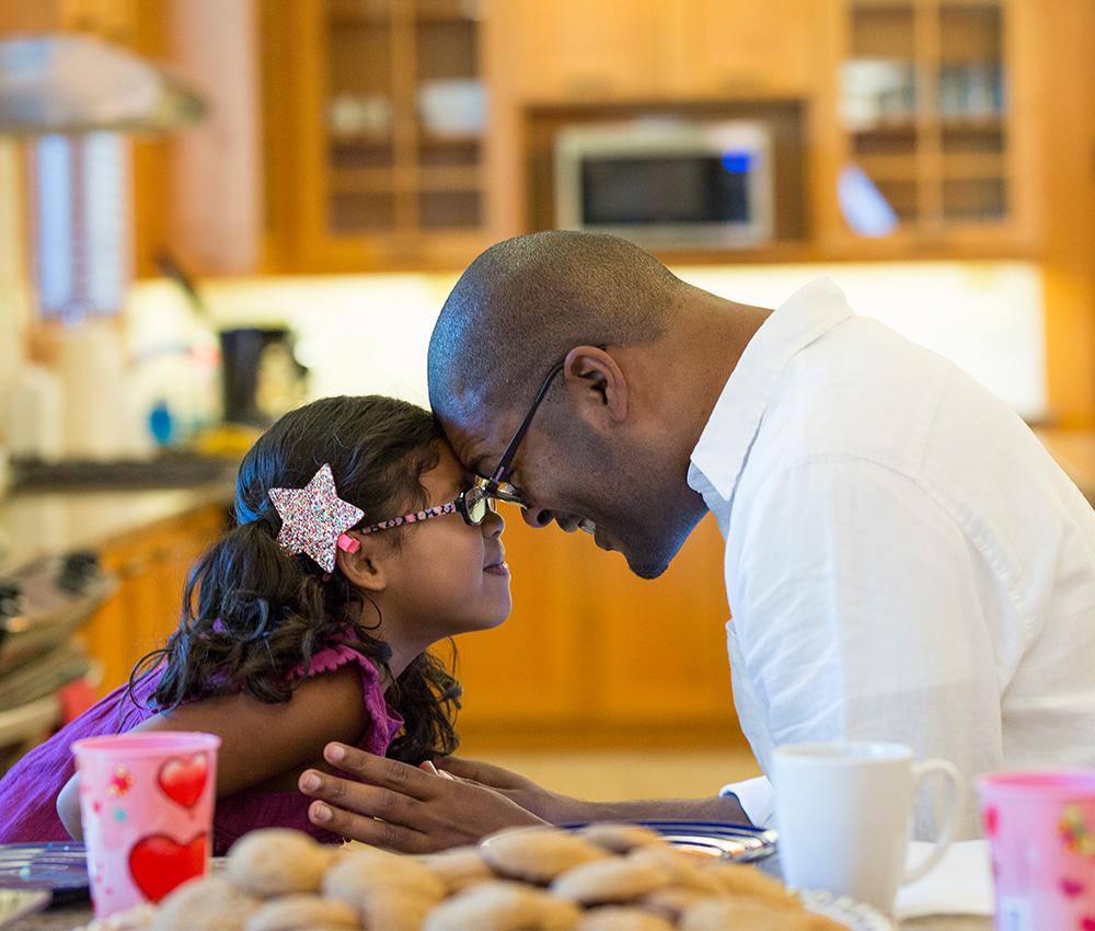 A father and daughter sit with their heads together in a cozy kitchen