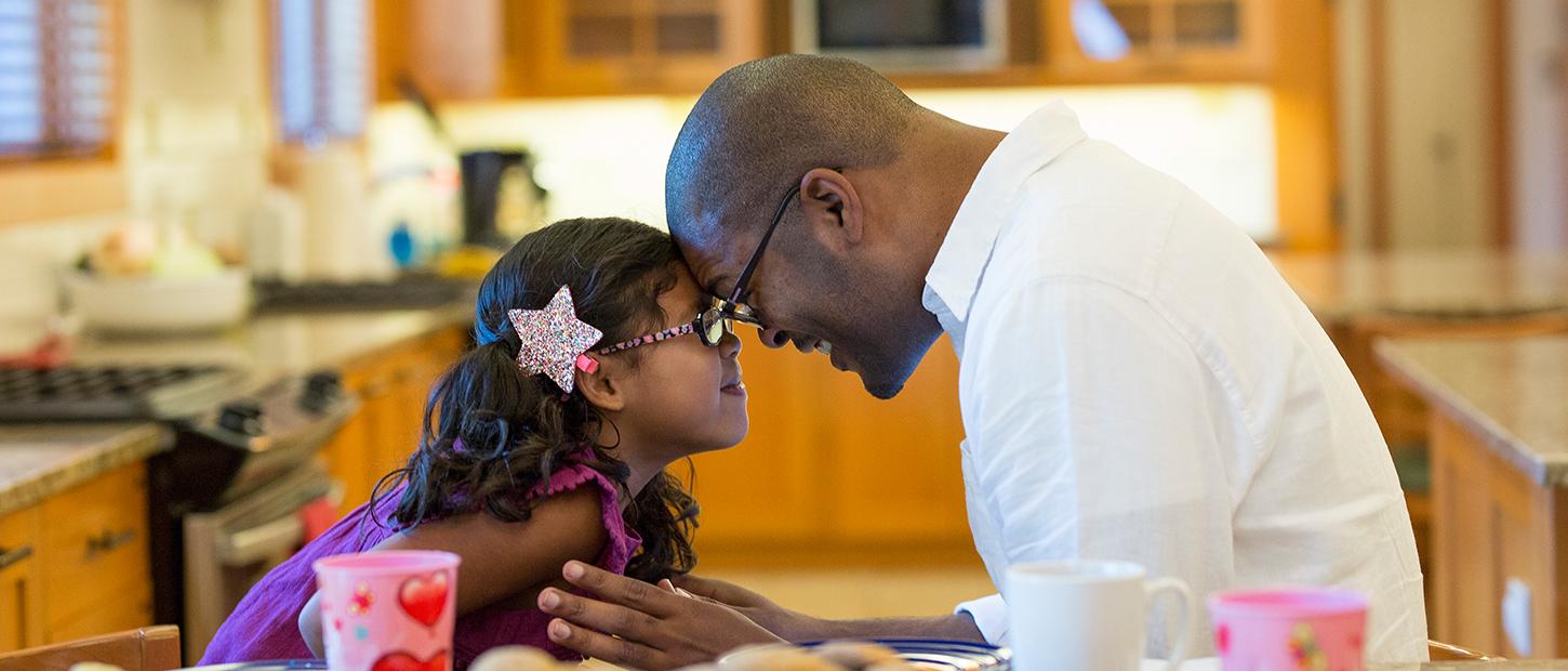 A father and daughter sit with their heads together in a cozy kitchen