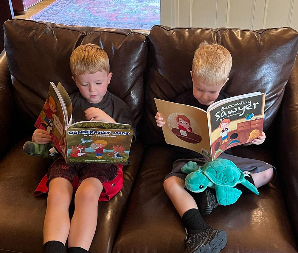 Two young boys sit together reading books on a big leather couch